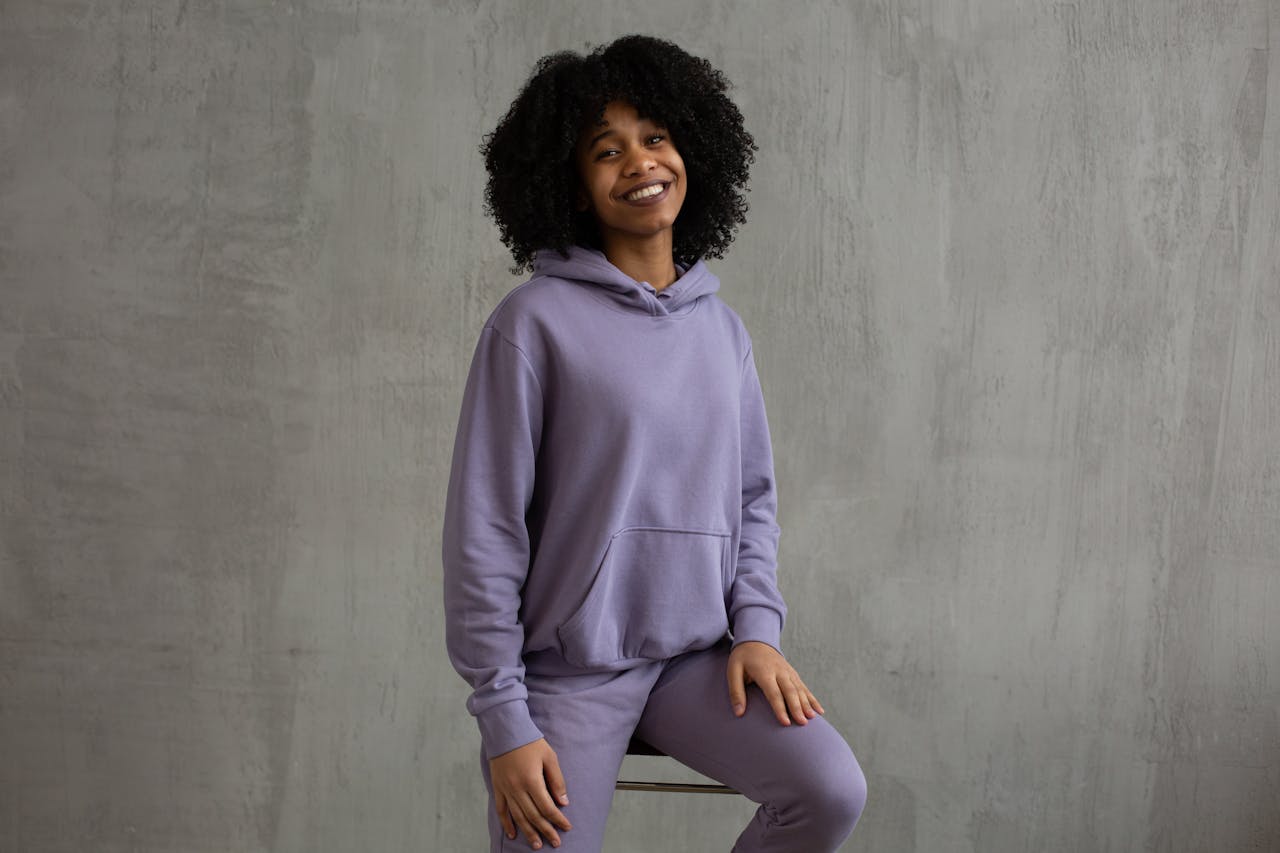 Joyful African American female with Afro hairstyle wearing violet hoodie and pants sitting on high chair against shabby gray wall and looking at camera with toothy smile